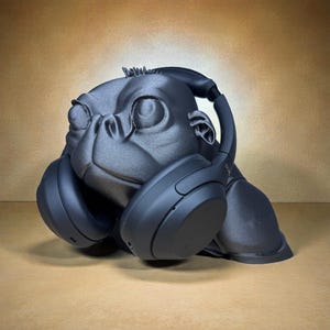 May include: A black gorilla-shaped headphone stand. The stand is designed to hold headphones, with the earcups resting on the gorilla's shoulders. The gorilla has a detailed face and is made of a matte black material.