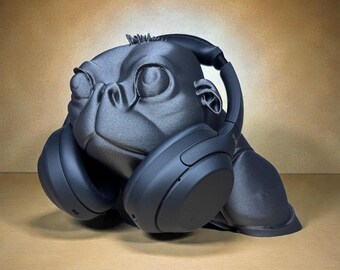 May include: A black gorilla-shaped headphone stand. The stand is designed to hold headphones, with the earcups resting on the gorilla's shoulders. The gorilla has a detailed face and is made of a matte black material.