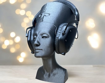 May include: A black bust of a pharaoh wearing black headphones. The bust is a dark gray color and has a detailed face and headdress. The headphones have large earcups and a padded headband.