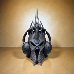 May include: A black headset stand shaped like a fantasy helmet with a blue gem and tall spikes. The stand is designed to hold a pair of black headphones. The stand is made of a dark gray material and sits on a light brown surface.