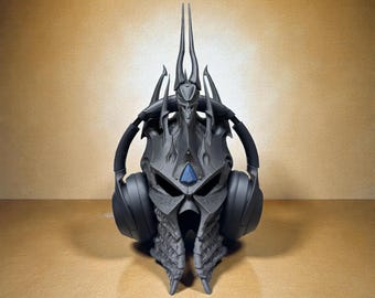 Puede incluir: Un soporte para auriculares negro con forma de casco de fantasía, con una gema azul y largas púas. El soporte está diseñado para sujetar un par de auriculares negros. El soporte está hecho de un material gris oscuro y se encuentra sobre una superficie marrón claro.