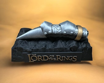 May include: A silver and gold metal replica of the One Ring from The Lord of the Rings, displayed on a black base with the words "The Lord of the Rings" inscribed on it.