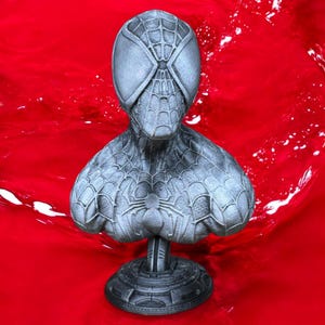 May include: A gray bust of Spider-Man, featuring the character's mask and torso. The bust has a detailed web pattern and a spider emblem on the chest. It is mounted on a circular base. The background is a vibrant red.
