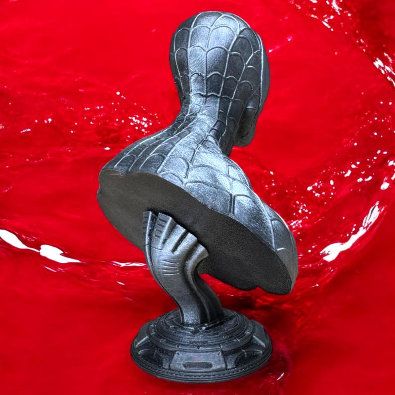 May include: A dark gray bust of a superhero with a spiderweb pattern on the head and shoulders. The bust is mounted on a decorative base and set against a vibrant red background.