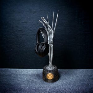 May include: A silver-colored headphone stand shaped like a skeletal hand holding a pair of black headphones. The base is dark gray with a gold-colored emblem. The stand is designed to hold headphones.