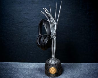 Puede incluir: Un soporte para auriculares de color plateado con forma de mano esquelética que sostiene unos auriculares negros. La base es de color gris oscuro con un emblema dorado. El soporte está diseñado para sujetar auriculares.
