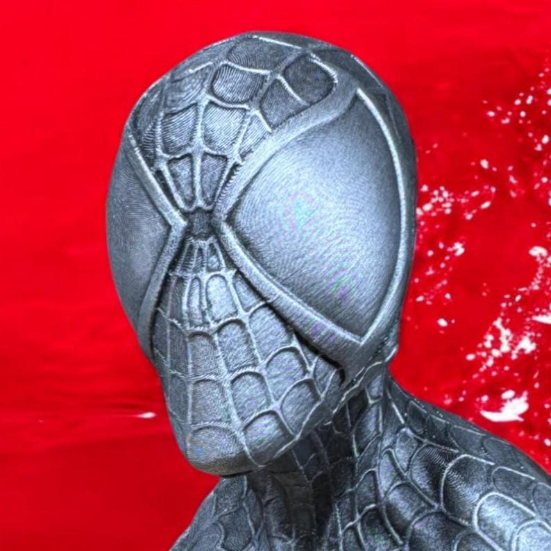 May include: A close-up of a black Spider-Man mask with a webbed design. The mask has large, oval eye openings and is set against a red background. The mask appears to be made of a textured material.