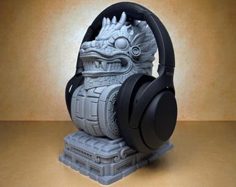 Puede incluir: Un soporte para auriculares gris impreso en 3D con forma de dios serpiente azteca. Los auriculares negros descansan sobre el soporte. El soporte tiene detalles intrincados y una superficie texturizada. El fondo es de color beige liso.