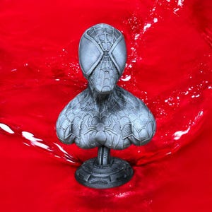 May include: A gray bust of Spider-Man, featuring the character's iconic mask and suit design. The bust is set against a vibrant red background, with a detailed spiderweb pattern across the surface.