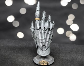Puede incluir: Una escultura de mano detallada de color plateado, al estilo de una armadura, con un anillo dorado en el dedo índice. La escultura está montada sobre una base redonda con la palabra "Sauron". El fondo es oscuro con luces blancas borrosas.