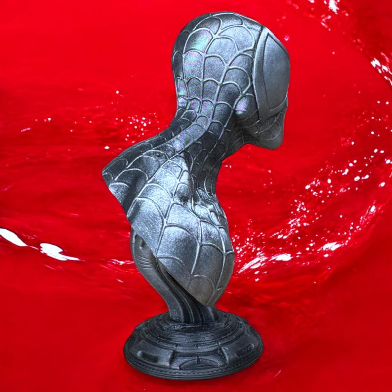 May include: A metallic gray bust of Spider-Man, featuring the character's mask and upper torso. The bust has a detailed web pattern and is set against a vibrant red background. The base is circular and also gray.
