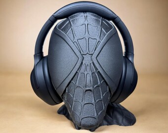 Puede incluir: Soporte para auriculares negro con temática de Spider-Man con diseño de telaraña. El soporte sostiene un par de auriculares negros supraaurales. El soporte está hecho de un material oscuro y tiene una apariencia tridimensional detallada.