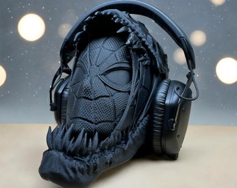 Puede incluir: Auriculares negros con un diseño impreso en 3D que presenta una máscara de Spider-Man y un conjunto de dientes. Los auriculares están sobre una superficie gris claro.