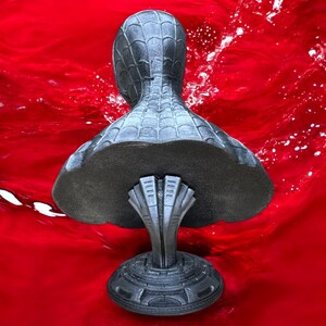 May include: A gray bust of a superhero, featuring a textured spiderweb pattern on the head and shoulders. The bust is mounted on a detailed pedestal with a stylized design. The background is a vibrant red.