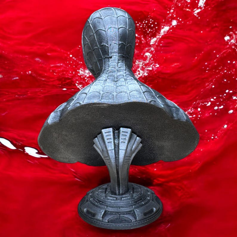 May include: A gray bust of a superhero, featuring a textured spiderweb pattern on the head and shoulders. The bust is mounted on a detailed pedestal with a stylized design. The background is a vibrant red.