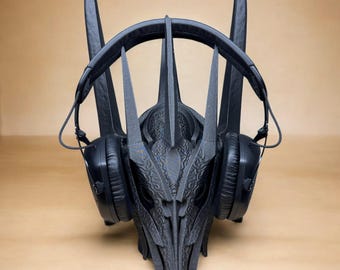 Puede incluir: Un soporte negro para auriculares con forma de cabeza de criatura con cuernos. El soporte tiene detalles intrincados y sostiene un par de auriculares negros supraaurales. El diseño es simétrico y se apoya sobre una superficie marrón claro.
