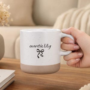 Puede incluir: Una taza de cerámica blanca con acabado moteado y base de color tostado. La taza presenta el texto negro "auntie lily" en letra cursiva sobre un pequeño gráfico de lazo negro. Tiene un asa curva en el lado derecho y un cuerpo cilíndrico.