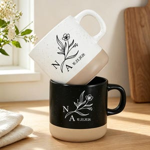 Puede incluir: Dos tazas de cerámica apiladas verticalmente, una blanca con motas negras y otra negra con motas blancas. Ambas presentan una ilustración floral, las letras N y A, y la fecha 10.23.2026. Cada taza tiene una base de color beige sólido.
