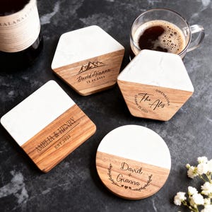 Peut inclure: Ensemble de dessous de verre personnalisés avec un design blanc et grain de bois. Chaque dessous de verre présente du texte et des dates personnalisés, certains incluant des graphiques de montagnes et des accents floraux. Une bouteille de vin et une tasse de café sont en arrière-plan.