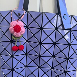 May include: A purple geometric tote bag with a crocheted flower and cherry keychain. The bag has a black strap and a black and white geometric pattern.