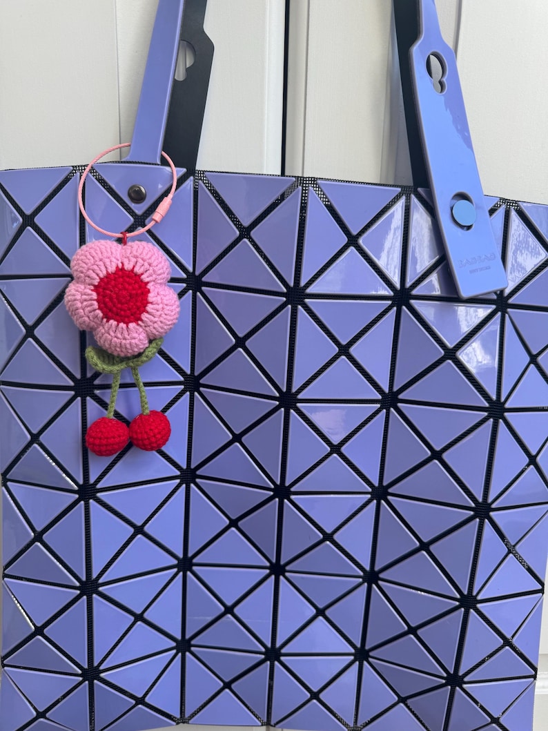 May include: A purple geometric tote bag with a crocheted flower and cherry keychain. The bag has a black strap and a black and white geometric pattern.
