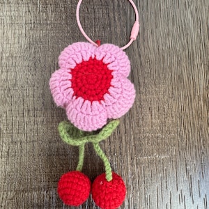May include: A pink and red crocheted flower with green stems and two red crocheted cherries hanging from the stems. The flower has a red center and a pink outer layer. The flower is attached to a pink and white string keychain.