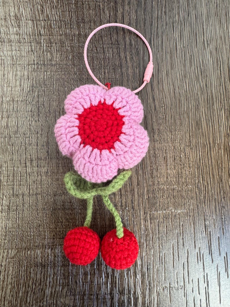 May include: A pink and red crocheted flower with green stems and two red crocheted cherries hanging from the stems. The flower has a red center and a pink outer layer. The flower is attached to a pink and white string keychain.