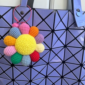 May include: A crocheted flower keychain in rainbow colors attached to a purple tote bag with a geometric pattern.