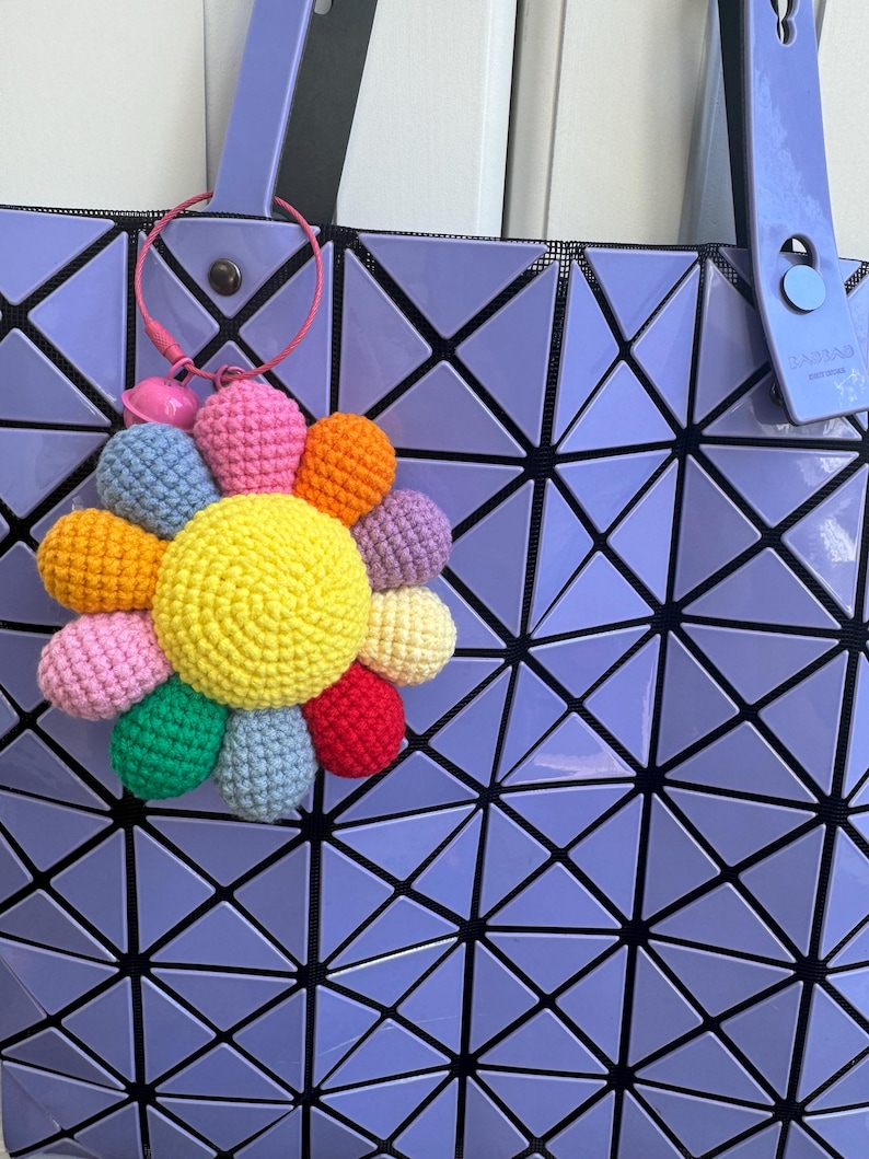 May include: A crocheted flower keychain in rainbow colors attached to a purple tote bag with a geometric pattern.