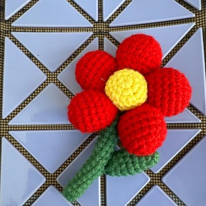 May include: A crocheted red flower with a yellow center and a green stem. The flower is made of individual crocheted circles that are sewn together.