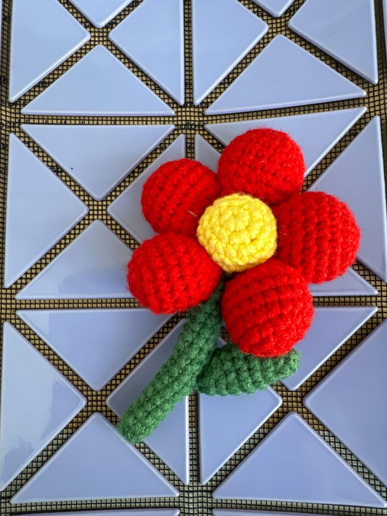 May include: A crocheted red flower with a yellow center and a green stem. The flower is made of individual crocheted circles that are sewn together.