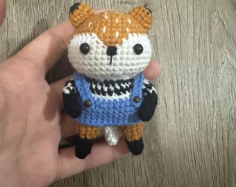 May include: A crocheted fox toy wearing a blue overall with white and black checkered trim. The fox has brown and white fur, black eyes, and a black nose.
