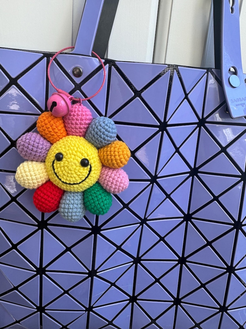 May include: A purple geometric tote bag with a crocheted rainbow flower keychain with a yellow smiley face. The keychain has a pink bell attached to it.