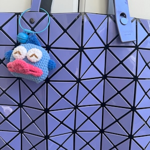 May include: A purple geometric tote bag with a black strap and a blue crocheted fish keychain. The bag has a geometric pattern of triangles.