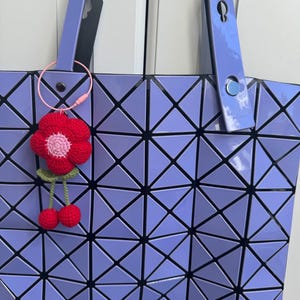 May include: A lavender tote bag with a geometric pattern. The bag has a black strap and a pink and red crocheted flower charm attached to the strap.