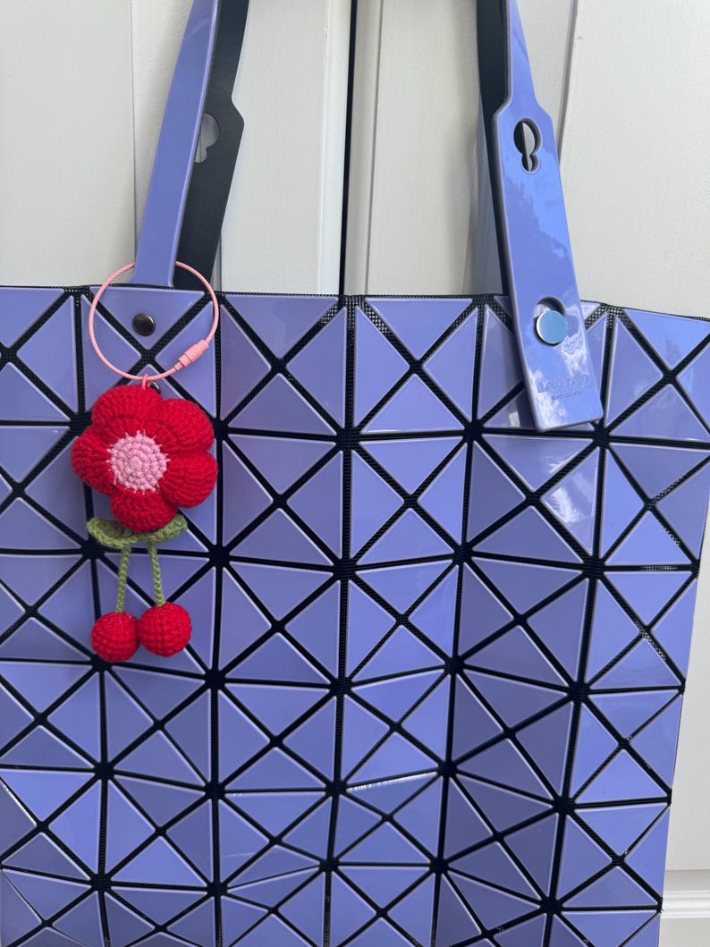 May include: A lavender tote bag with a geometric pattern. The bag has a black strap and a pink and red crocheted flower charm attached to the strap.