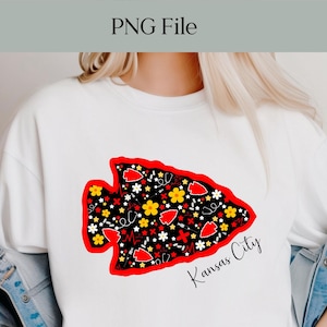 May include: White t-shirt featuring a large, stylized arrowhead design with a black background and red outline. The arrowhead is filled with red hearts, yellow flowers, and white stars. The words "Kansas City" are written in cursive below the arrowhead.