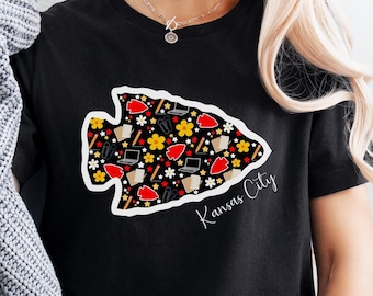 May include: Black t-shirt with a white arrowhead design filled with red, yellow, and black illustrations. The text "Kansas City" is written in white below the arrowhead.