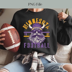 Pode incluir: Camisola preta com um gráfico de um Viking com barba roxa, óculos de sol e chapéu com chifres. O texto "MINNESOTA FOOTBALL 1961" é impresso em amarelo e roxo. Uma bola de futebol castanha é segurada na mão esquerda.