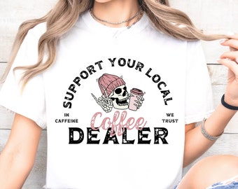 May include: White t-shirt with a black and white graphic design featuring a skull wearing a pink beanie and holding a coffee cup. The text reads "Support Your Local In Caffeine We Trust Coffee Dealer".