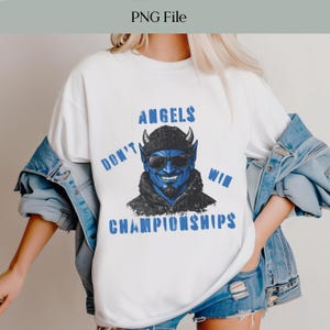 May include: White t-shirt with a blue graphic of a devil wearing sunglasses and a beanie. The text "DON'T ANGELS WIN CHAMPIONSHIPS" is printed in blue. The shirt is worn by a person wearing a denim jacket.