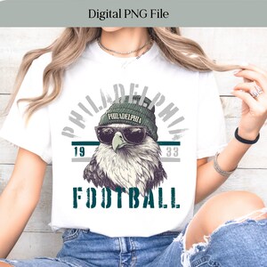 Può includere: T-shirt bianca con una grafica di un'aquila con occhiali da sole e berretto. Il design include le scritte "PHILADELPHIA" e "FOOTBALL", insieme ai numeri "19" e "33". Le parole "Digital PNG File" sono in alto.