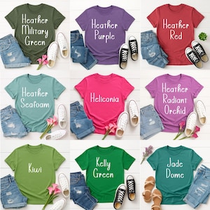 May include: A collection of colorful t-shirts in various shades, including green, purple, and red, each paired with denim shorts and sneakers. The shirts feature text such as "Heather Military Green" and "Heliconia". A stylish and casual clothing ensemble.