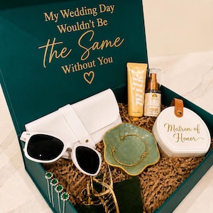 May include: A dark green gift box with the text My Wedding Day Wouldn't Be the Same Without You. Inside are white sunglasses, a white pouch, a gold hair clip, green hair pins, a gold bracelet in a green dish, a tube of lotion, and a white case labeled Matron of Honor.