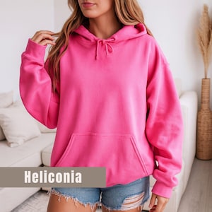 Op de afbeelding: Een felroze hoodie met een capuchon met trekkoord en een kangoeroezak. Het woord "Heliconia" wordt weergegeven in een rechthoekige banner onderaan de afbeelding. De hoodie wordt gecombineerd met een spijkerbroek.