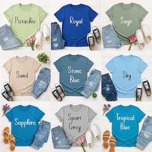 May include: A collection of t-shirts in various colors, including pistachio, royal blue, sage, sand, stone blue, sky, sapphire, sport grey, and tropical blue. Each shirt is paired with denim shorts and sneakers or sandals. The shirts have text on them.