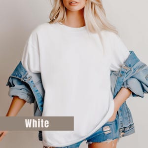 May include: A white, oversized t-shirt with the word "White" in a brown rectangle at the bottom. The shirt is paired with a light blue denim jacket and denim shorts. The model has blonde hair.