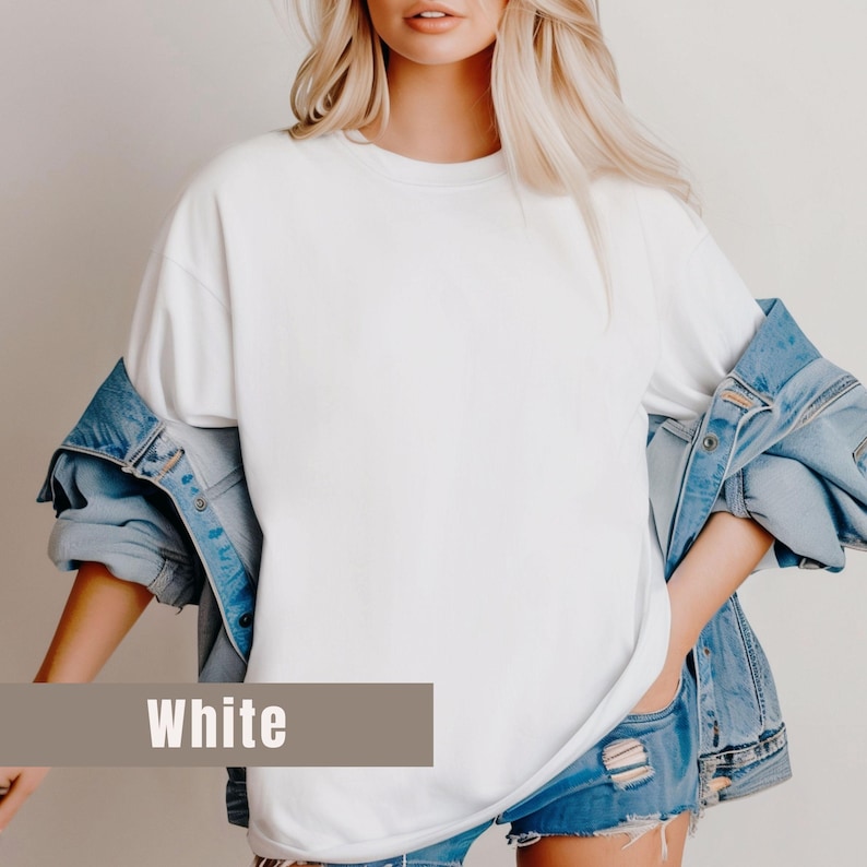May include: A white, oversized t-shirt with the word "White" in a brown rectangle at the bottom. The shirt is paired with a light blue denim jacket and denim shorts. The model has blonde hair.
