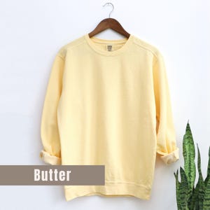May include: A pale yellow sweatshirt with long sleeves, displayed on a wooden hanger against a white background. The word "Butter" is printed on a rectangular label at the bottom. The sleeves are cuffed at the wrists.