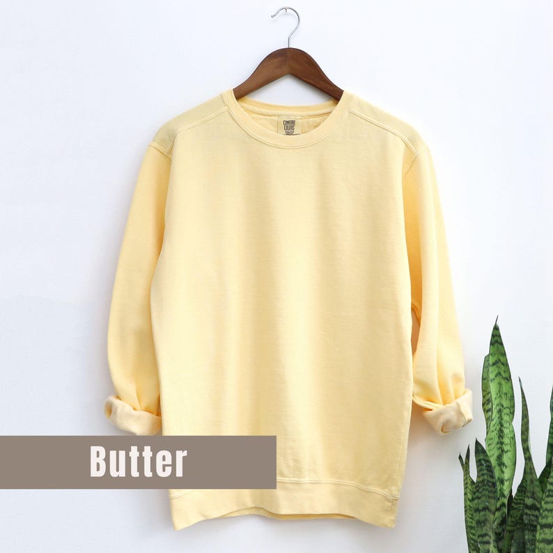 May include: A pale yellow sweatshirt with long sleeves, displayed on a wooden hanger against a white background. The word "Butter" is printed on a rectangular label at the bottom. The sleeves are cuffed at the wrists.